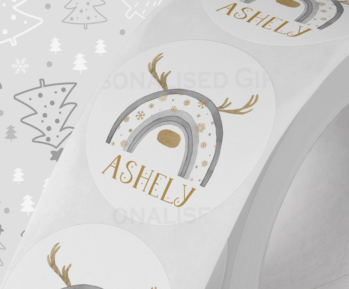Glossy white round Christmas sticker personalised with a name, printed with a grey rainbow reindeer design, shown on a strip of stickers