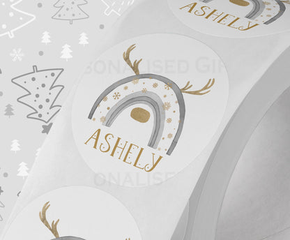 Glossy white round Christmas sticker personalised with a name, printed with a grey rainbow reindeer design, shown on a strip of stickers