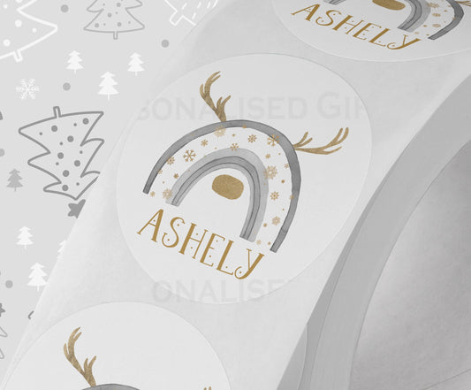 Glossy white round Christmas sticker personalised with a name, printed with a grey rainbow reindeer design, shown on a strip of stickers