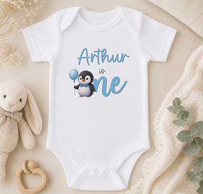 A white onesie with a penguin graphic and the text "Arthur is one" printed on it, along with a stuffed animal and some decorative elements.