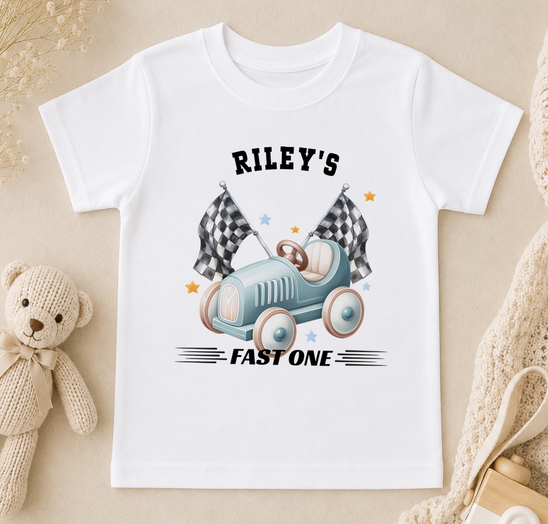 Personalised white t-shirt printed with a blue toy racing car, chequered flags and 'Riley's FAST ONE'.
