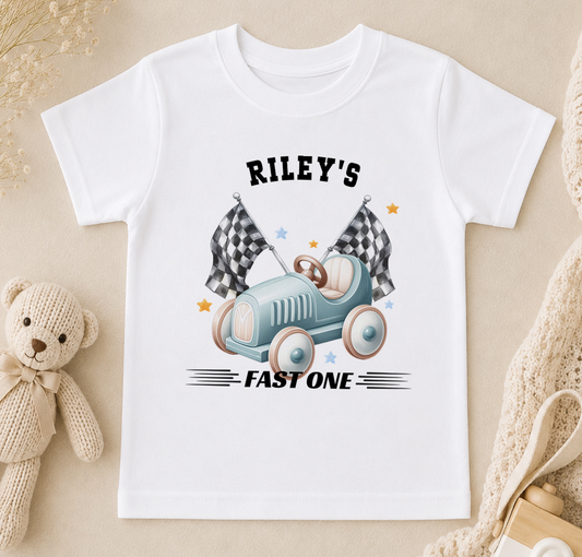 A white t-shirt with a graphic design of a blue toy car with checkered flags and the text "Riley's FAST ONE" printed on it.