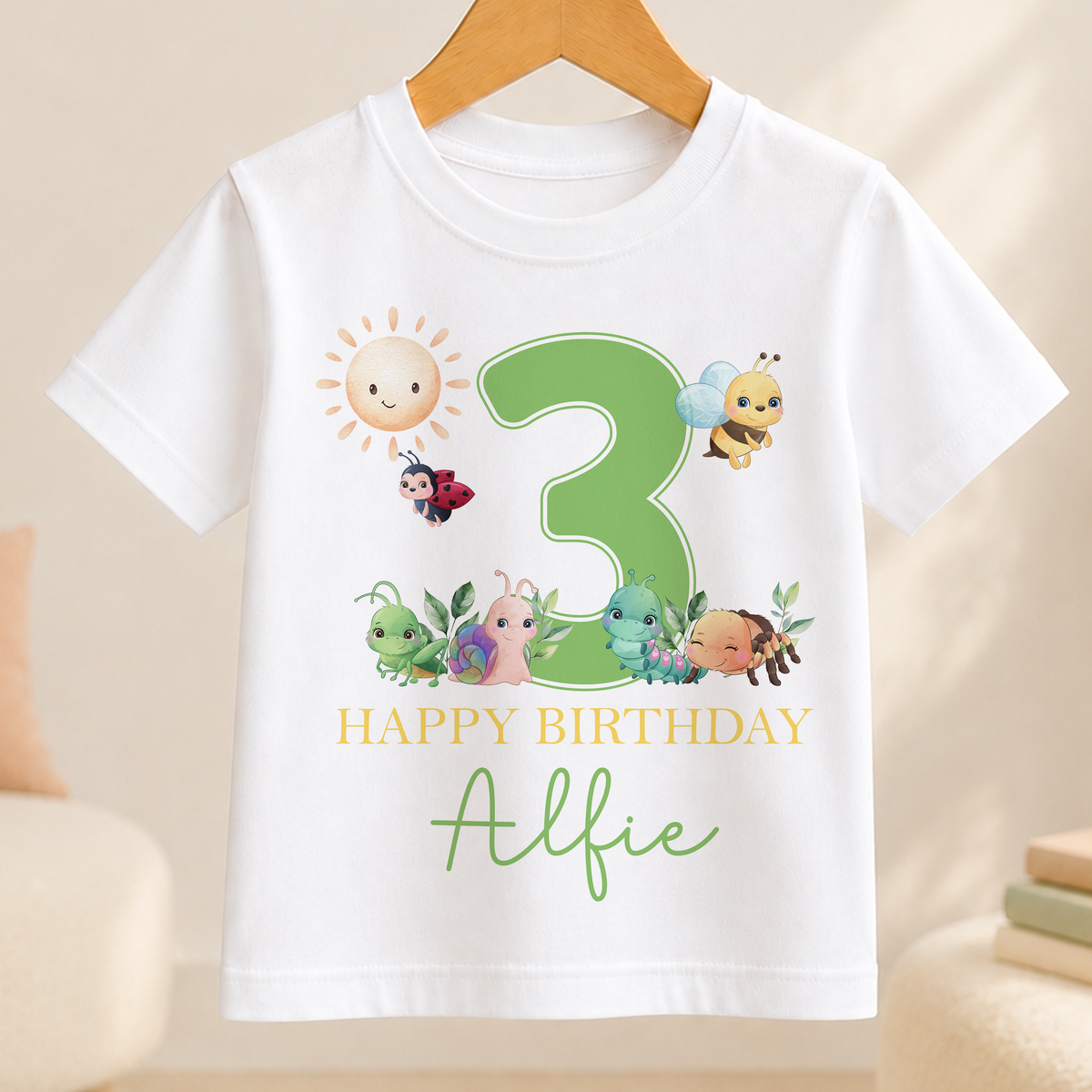 A white t-shirt with a green number 3 and various cartoon insects, including a ladybug, on it. The text below the number reads "Happy Birthday Alfie".