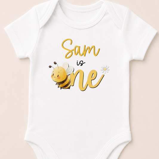 A white baby onesie with a yellow bee and the text "Sam is one" printed on it.