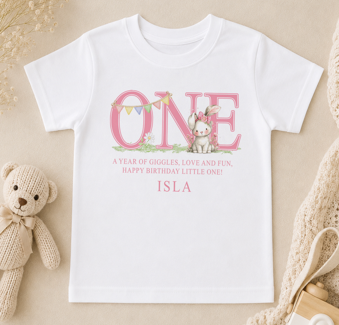A white t-shirt with a pink text design that says "ONE" and features a small rabbit character. Below the text, there is a message that reads "A year of giggles, love and fun, happy birthday little one! ISLA".