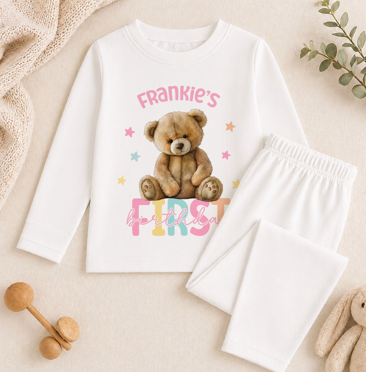 A white long-sleeved shirt with a teddy bear graphic and the text "Frankie's first birthday" printed on it, along with a pair of white pajama pants.