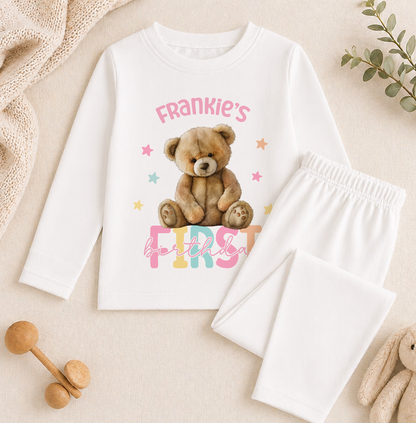 A white long-sleeved shirt with a teddy bear graphic and the text "Frankie's first birthday" printed on it, along with a pair of white pajama pants.