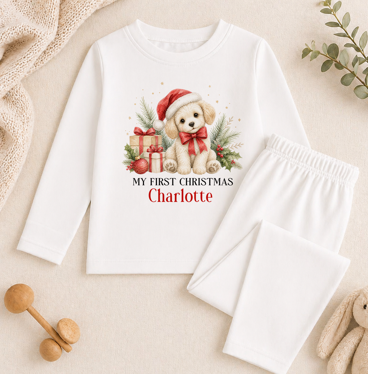 A white long-sleeved shirt with a Christmas-themed graphic of a dog wearing a Santa hat and holding a gift, along with the text "My first Christmas Charlotte". The shirt is displayed on a beige background, and there are some Christmas-themed items such as a teddy bear and a toy car nearby.