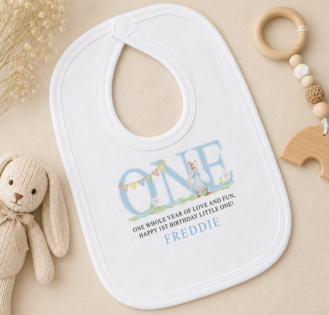 A white baby bib with a personalized message, a wooden bead rattle, and a stuffed animal rabbit on a beige surface.