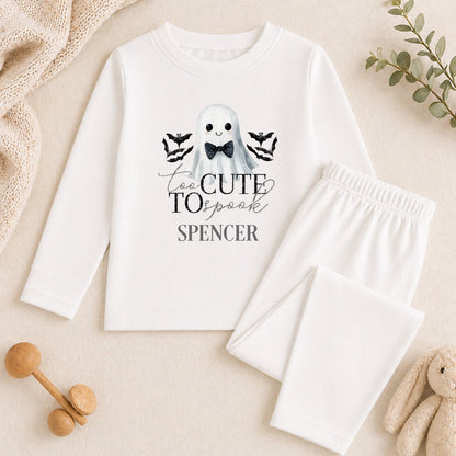 A white long-sleeved shirt with a graphic of a ghost and the text "Too Cute to Spank" printed on it, along with a pair of white pajama pants. The shirt and pants are placed on a beige surface, and there are some toys and greenery in the background.