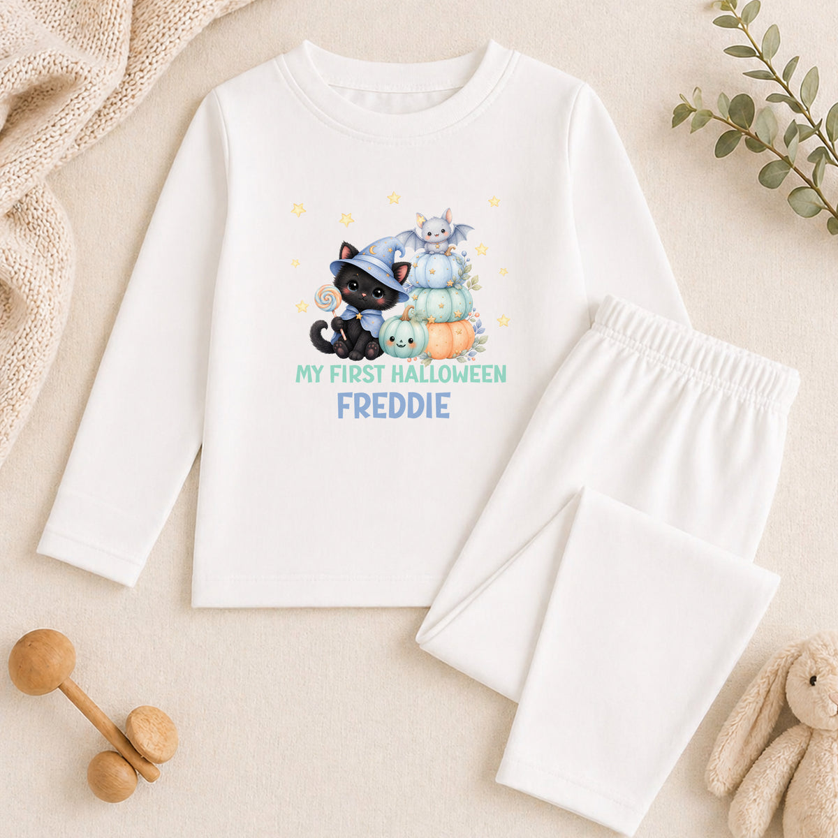 A white long-sleeved shirt with a Halloween-themed graphic and text, along with a pair of white pajama pants, and some stuffed animals on a beige surface.