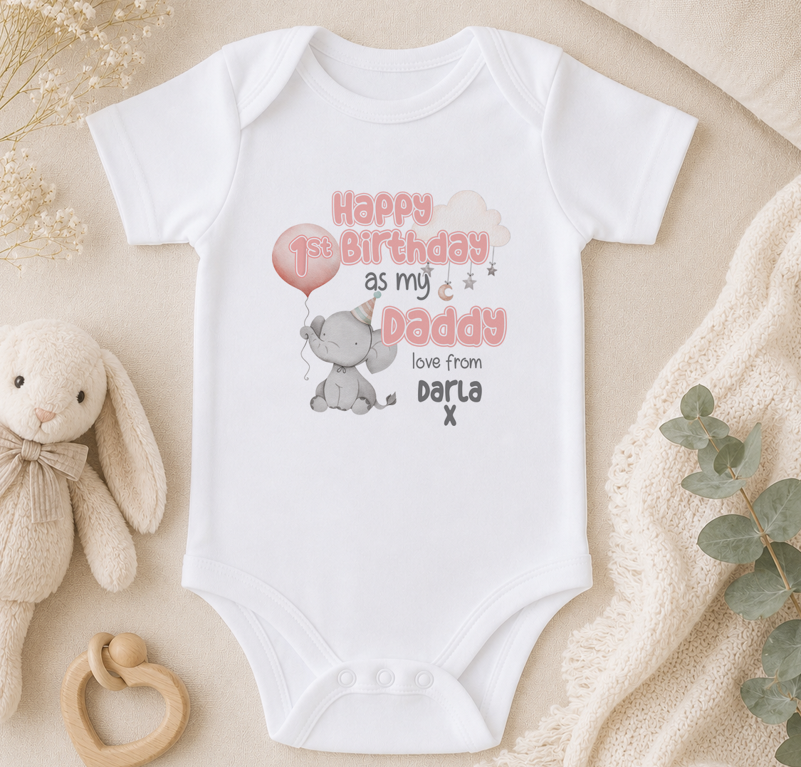 A white onesie with a birthday message and an elephant graphic, along with a stuffed animal and some greenery, is displayed on a surface.