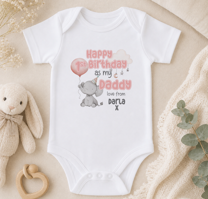 A white onesie with a birthday message and an elephant graphic, along with a stuffed animal and some greenery, is displayed on a surface.