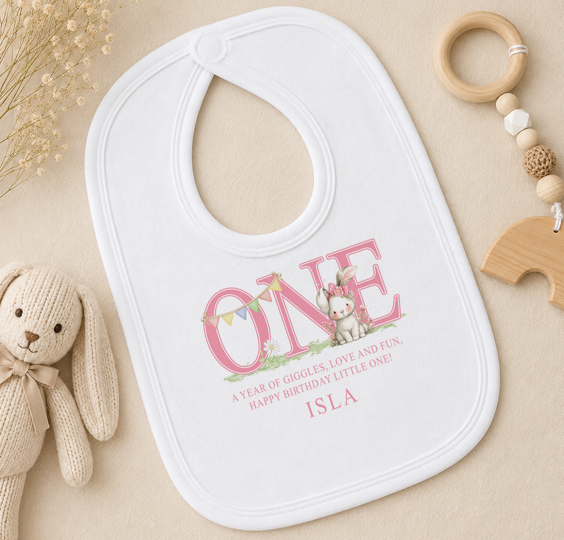 A white bib with a personalized message, a wooden toy, and some dried flowers on a beige surface.