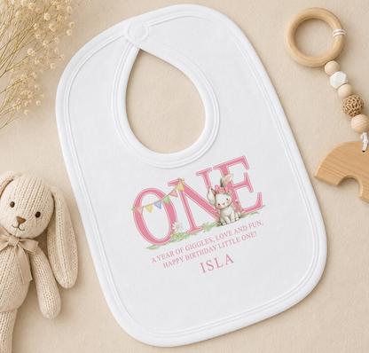 A white bib with a personalized message, a wooden toy, and some dried flowers on a beige surface.