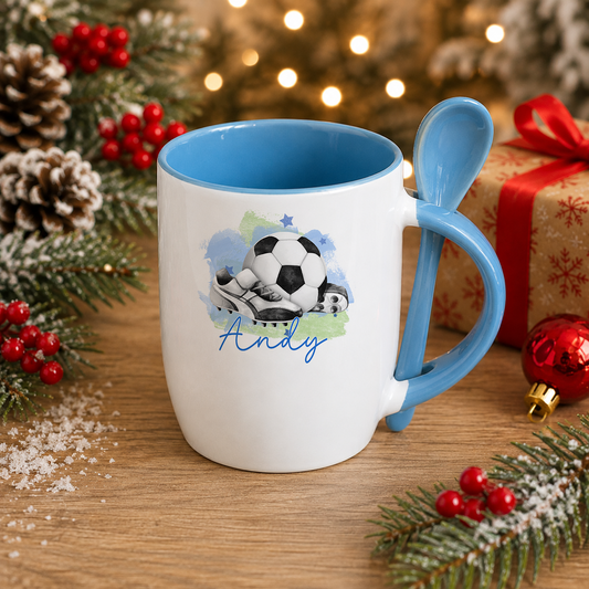 A white mug with a blue handle and a soccer ball design sits on a wooden surface, surrounded by Christmas decorations and a gift box.