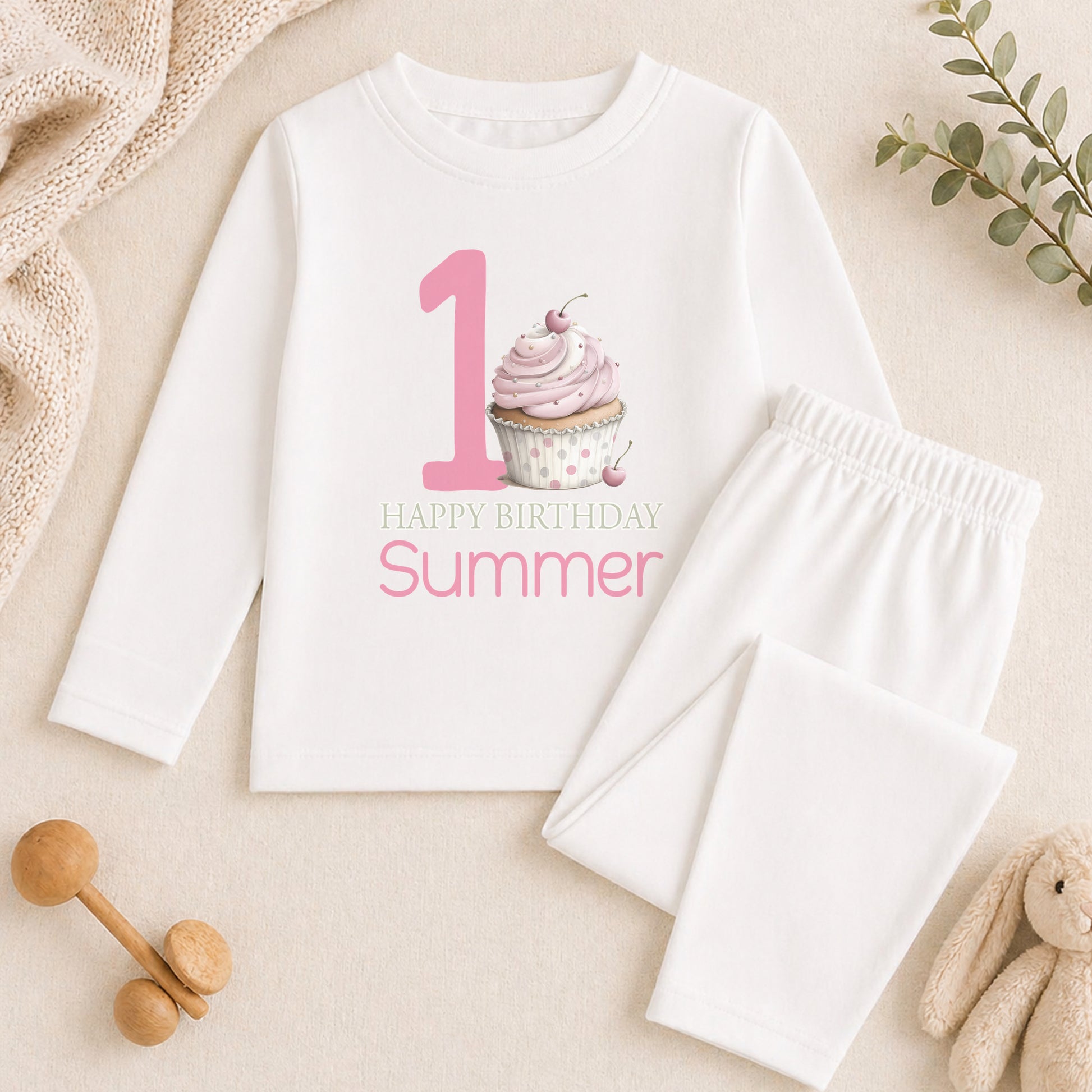 A white long-sleeved shirt with a graphic of a cupcake and the text "Happy Birthday Summer" printed on it, along with a pair of white pajama pants. The shirt and pants are placed on a beige surface, and there are some toys and a plant in the background.