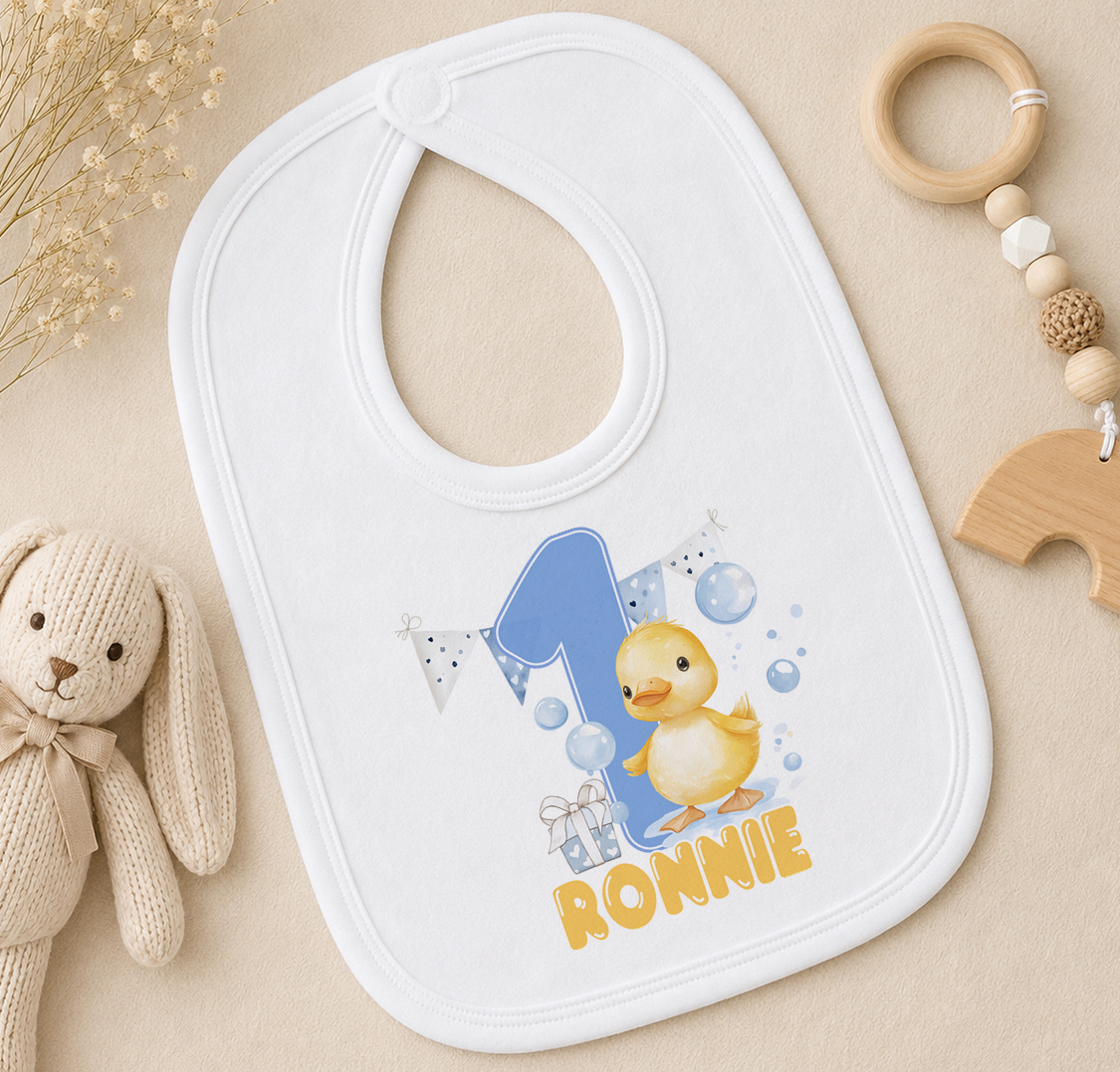 A white bib with a cartoon duck and the number 1, along with the name "Ronnie" printed on it, is placed on a surface next to a stuffed animal.