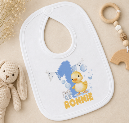 A white bib with a cartoon duck and the number 1, along with the name "Ronnie" printed on it, is placed on a surface next to a stuffed animal.