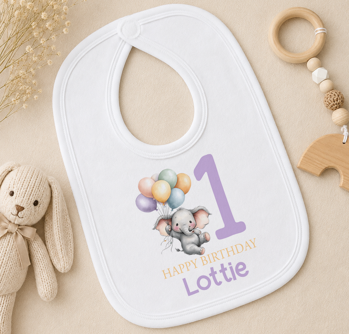 A white baby bib with a cartoon elephant and balloons, along with a wooden toy and a wooden bead ring, are arranged on a beige surface.
