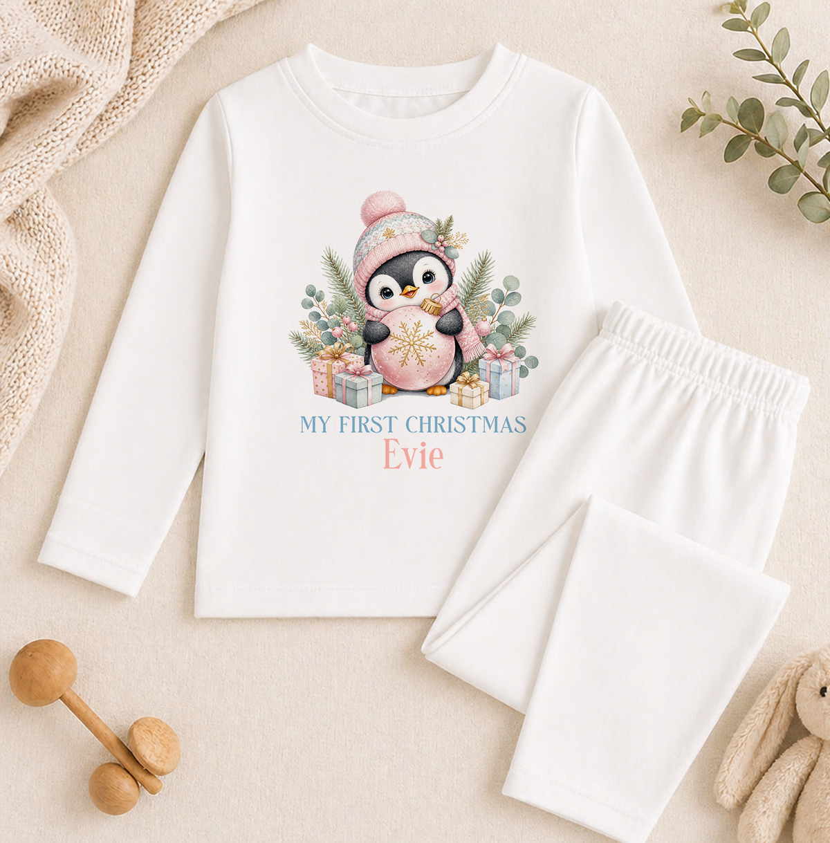 A white long-sleeved shirt with a penguin graphic and the text "My first christmas Eve" printed on it, along with a pair of white pajama pants. The shirt and pants are placed on a beige background, and there are some wooden toys and a plant visible in the image.