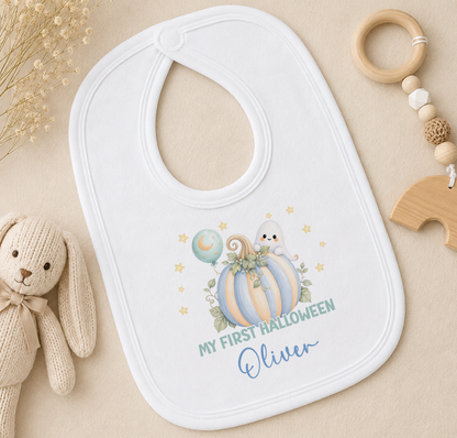 A white baby bib with a pumpkin and ghost design, along with the text "My first Halloween Oliver" printed on it, is placed on a beige surface next to a stuffed rabbit toy and a wooden bead ringer.