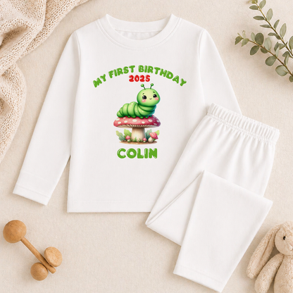 A white long-sleeved shirt with a printed design of a caterpillar on a mushroom, along with the text "My first birthday 2015 COLIN" printed on it. The shirt is placed on a beige surface, and there are some toys and a plant in the background.