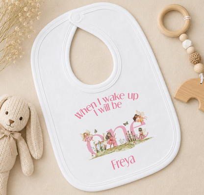 White baby bib printed with fairy illustrations, styled with a wooden toy and a teething ring