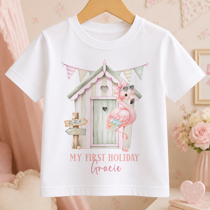 A white t-shirt with a pink flamingo and a beach house graphic, along with the text "My first holiday Gracie".
