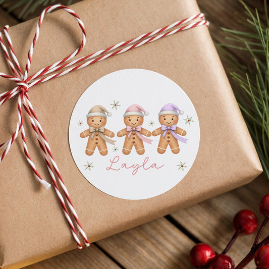 Round white Christmas gift label sticker featuring three smiling gingerbread girls in pink, purple and yellow hats, personalised with a custom name, on a brown gift box with red and white striped ribbon