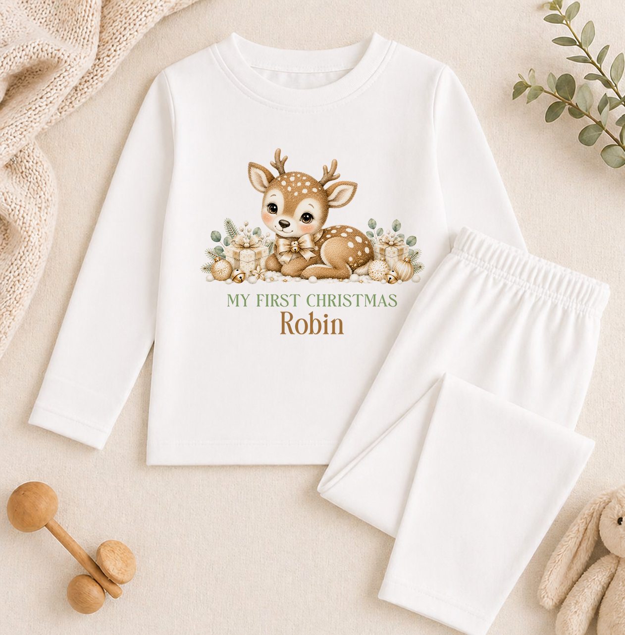 A white long-sleeved shirt with a cartoon deer and the text "My first Christmas Robin" printed on it, along with a pair of white pajama pants. The shirt and pants are placed on a beige background, and there are some wooden toys and a plant visible in the image.