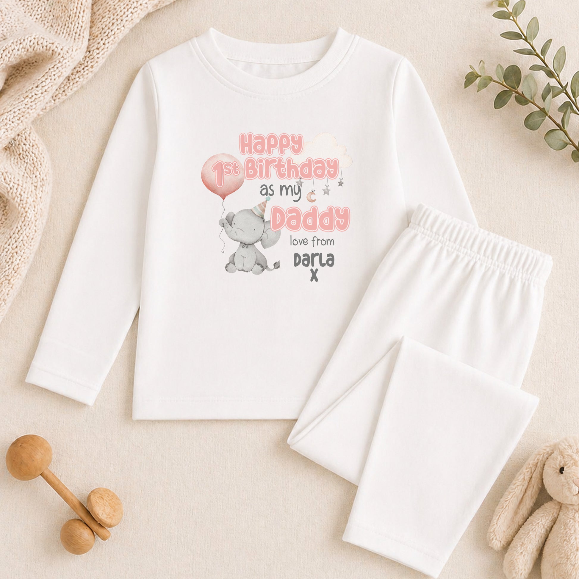 A white long-sleeved shirt with a birthday message and a cute elephant graphic, along with a pair of white pajama pants, a stuffed animal, and some wooden toys.