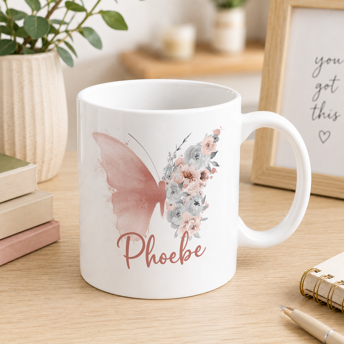 A white ceramic mug with a floral design and the name "Phoebe" printed on it, placed on a wooden surface next to some books and a plant.
