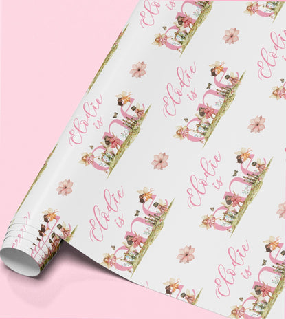Roll of pink floral fairy wrapping paper printed with 'Ella is' as part of the personalised first birthday design.
