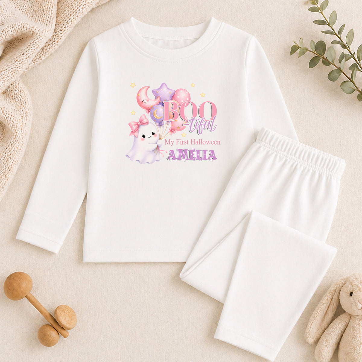 A white long-sleeved shirt with a Halloween-themed graphic, a pair of white pajama pants, and some stuffed animals on a beige background.