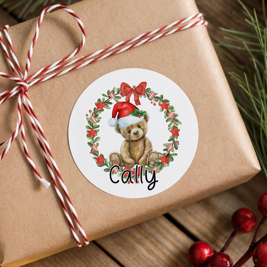 Round white Christmas sticker featuring a teddy bear wreath design with a red Santa hat and green bow, personalised with the custom name Cally, on a brown gift box with red and white striped ribbon