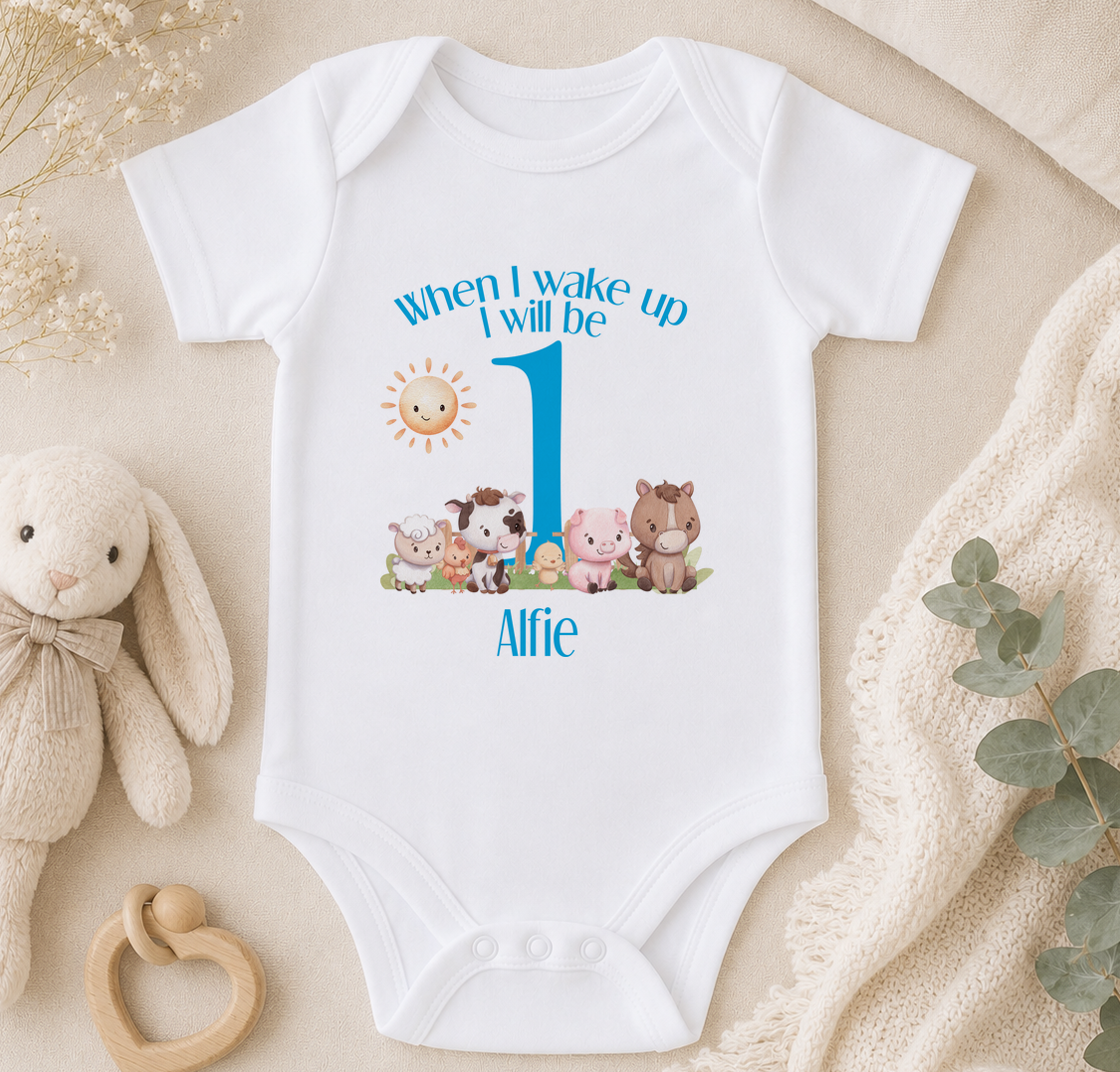 A white onesie with a cartoon image of farm animals and the text "When I wake up I will be 1" printed on it, along with the name "Alfie". The onesie is placed on a beige surface, surrounded by a stuffed animal, a teething ring, and some greenery.