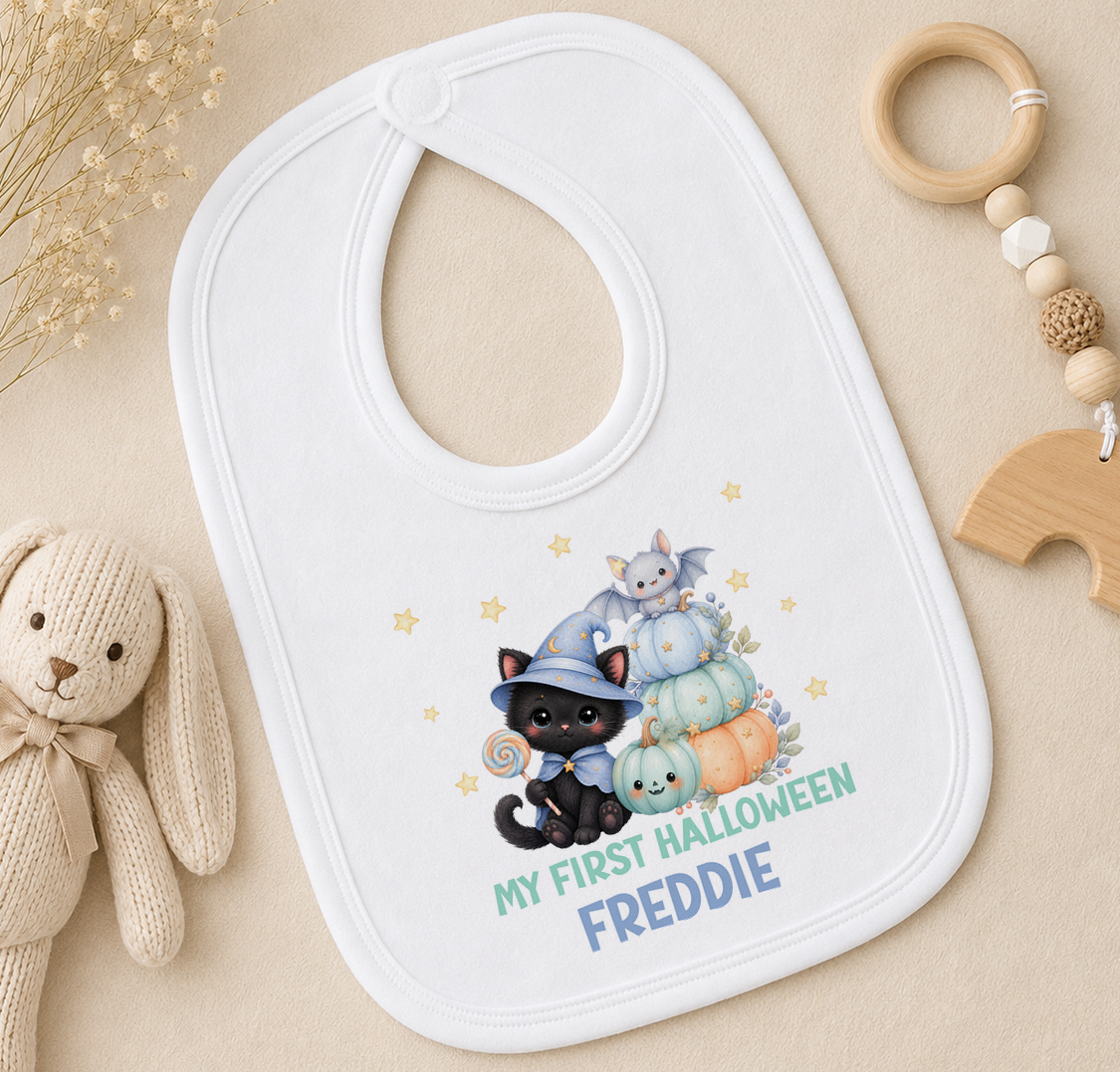 A white baby bib with a Halloween-themed design, featuring a black cat, pumpkins, and stars, along with the text "My first HALLOWEEN FEDDIE".