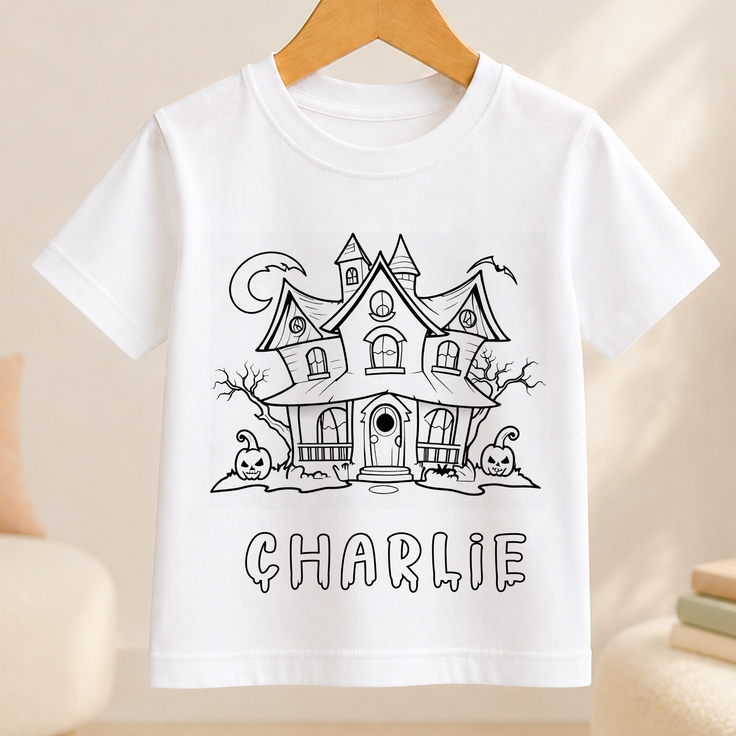 A white t-shirt with a black and white illustration of a house with a castle-like appearance, featuring a crescent moon and two jack-o'-lanterns. The name "CHARLIE" is written below the illustration.
