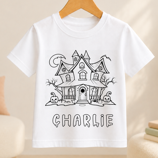 A white t-shirt with a black and white illustration of a house with a castle-like appearance, featuring a crescent moon and two jack-o'-lanterns. The name "CHARLIE" is written below the illustration.