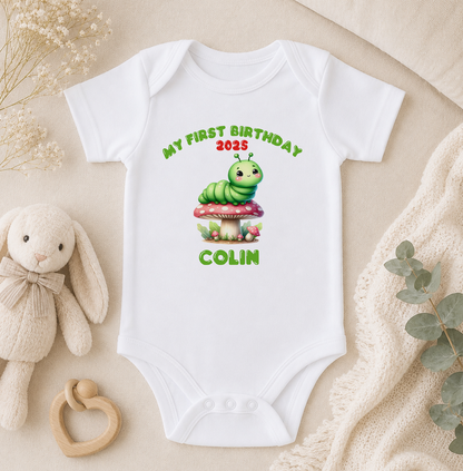 A white baby onesie with a green caterpillar graphic and text, along with a stuffed rabbit and a wooden bead rattle, all arranged on a beige surface.