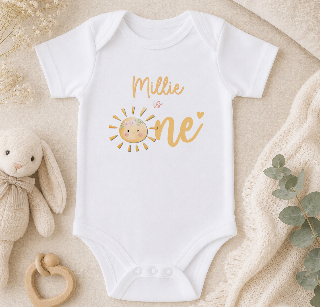 A white onesie with a sun graphic and the text "Milly is one" printed on it, along with a cute bunny plush toy and some greenery.