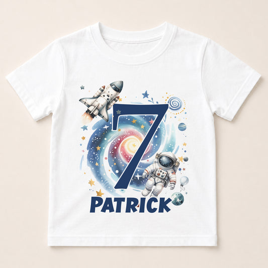 A white t-shirt with a space-themed design featuring a rocket ship, an astronaut, and a spiral galaxy. The number 7 is prominently displayed in the center, and the name "PATRICK" is written below the number.