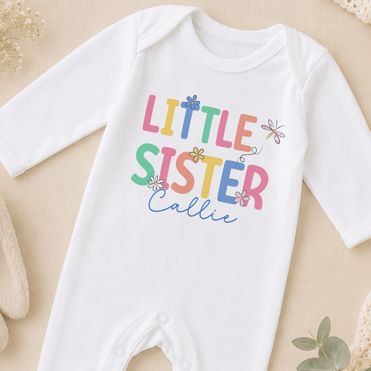 A white onesie with the text "LITTLE SISTER" printed on it, along with colorful flowers and a dragonfly design.
