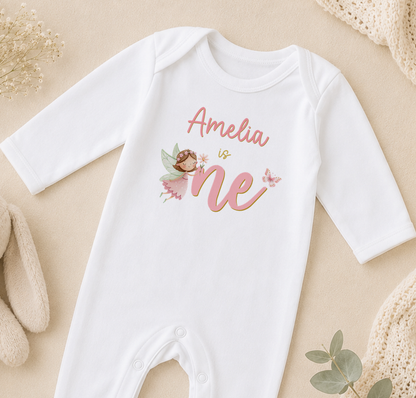 A white onesie with a personalized name and a fairy design, placed on a beige surface with some baby items and a plant in the background.