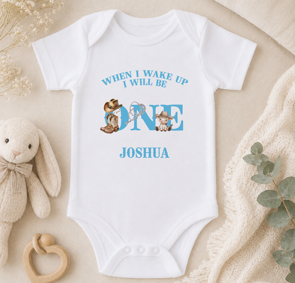 A white onesie with a blue text design that says "WHEN I WAKE UP I WILL BE ONE JOSHUA" and features a cartoon image of a baby rabbit.