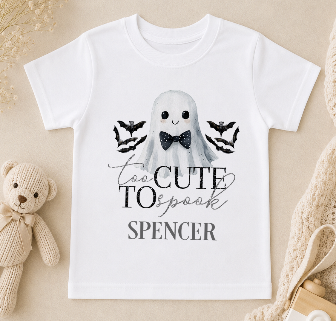 A white t-shirt with a graphic design featuring a ghost character wearing a bow tie and surrounded by bats. The text on the t-shirt says "Too Cute to Spook Spencer".