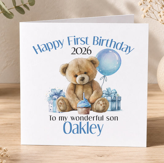 White first birthday card featuring a blue teddy bear, a cupcake and gift boxes, printed with 'Happy First Birthday 2026 To my wonderful son Oakley' as a personalisation example.