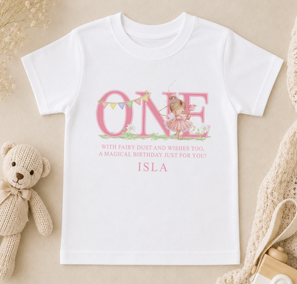 A white t-shirt with a pink design and text, along with a teddy bear and other baby items, arranged on a beige surface.