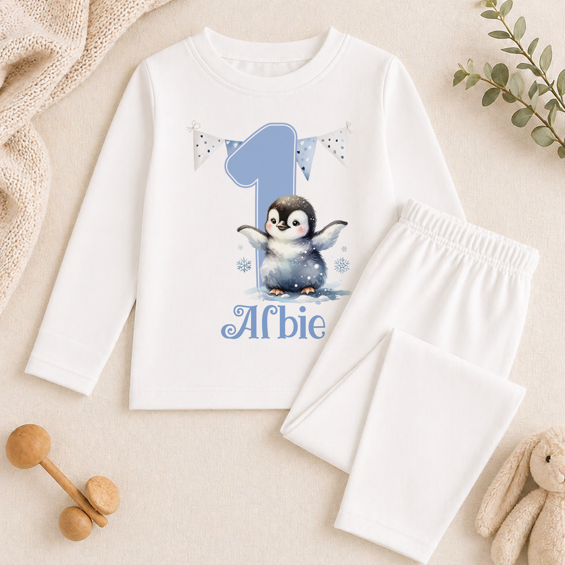 A white long-sleeved shirt with a penguin design and the number 1, along with a pair of white pants, are displayed on a beige surface. There are also some stuffed animals and a plant in the background.